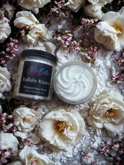 Two jars of whipped body butter labeled 'Lullaby Kisses' surrounded by white flowers on a textured surface.